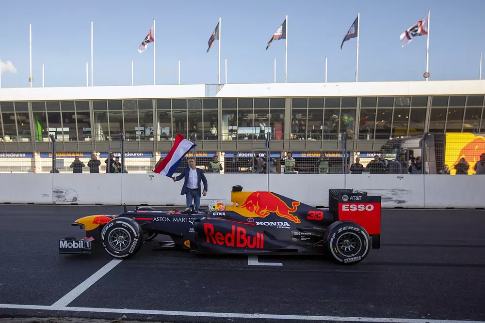 Max Verstappen, RB8, op start/finish van Circuit Zandvoort met Jan Lammers op de achtergrond die met de Nederlandse vlag zwaait
