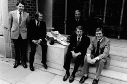 John Barnard, Ron Dennis en Teddy Mayer achter Tyler Alexander en Creighton Brown poseren met een mo