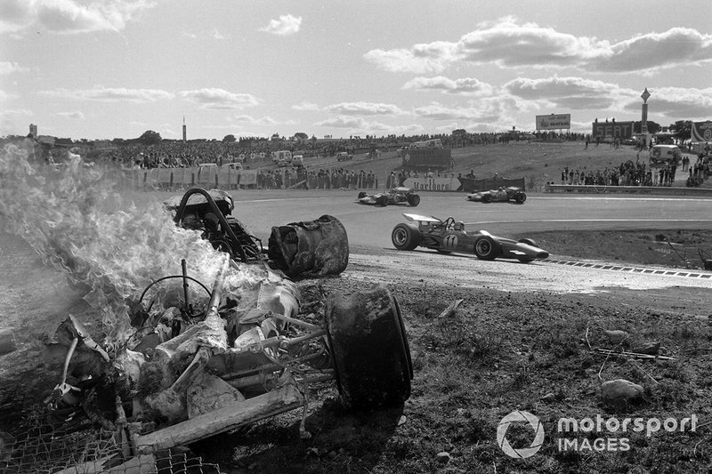 Bruce McLaren, McLaren M14A Ford, Graham Hill, Lotus 49C Ford, y Rolf Stommelen, Brabham BT33 Ford, pasan un un coche en llamas