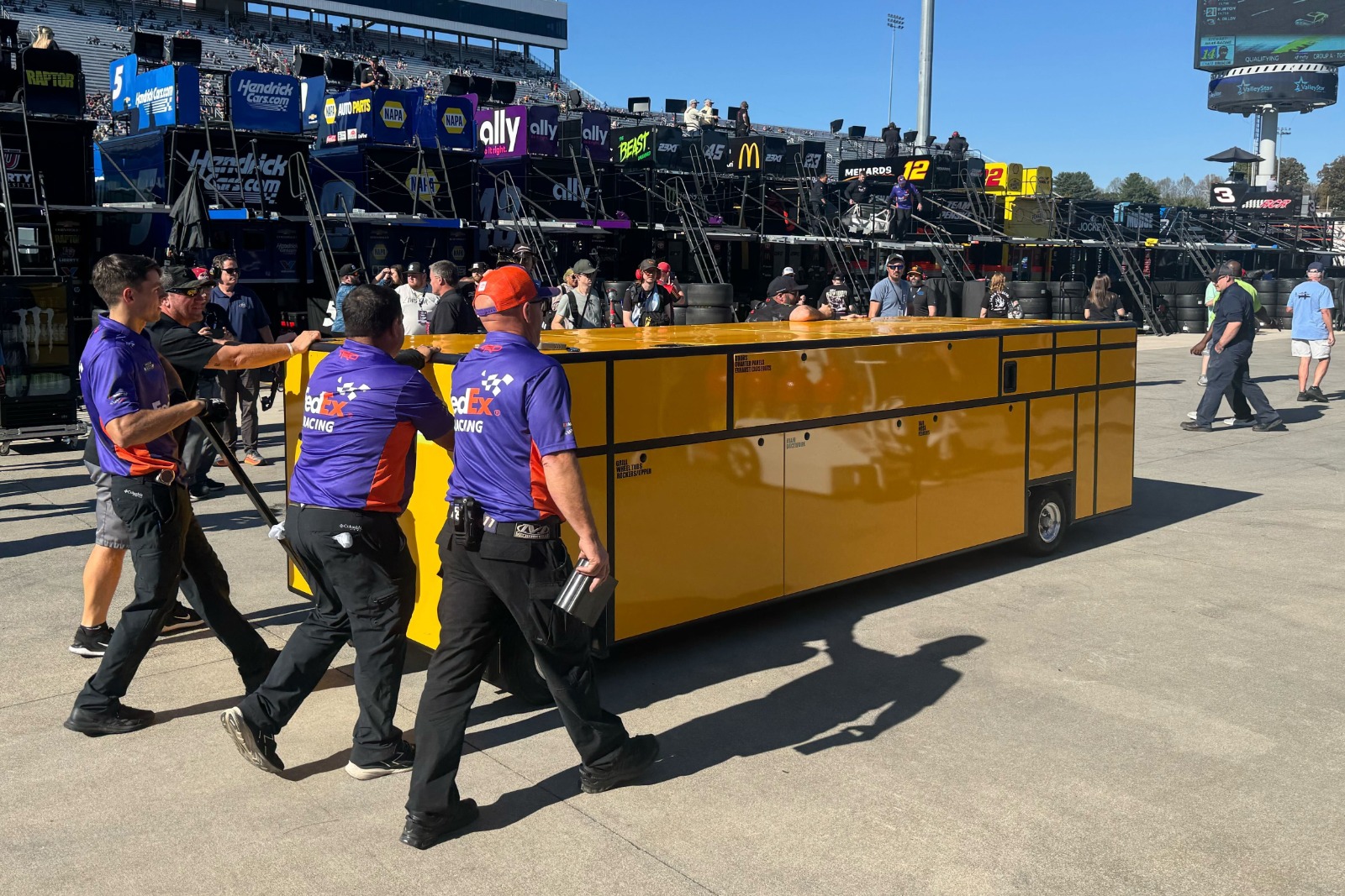 The crew of the No. 11 car walks a yellow compartment box on wheels to their garage.